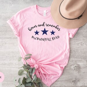 以下が含まれることがあります： 「honor and remember MEMORIAL DAY」という文字と3つの青い星がプリントされたピンクのTシャツです。