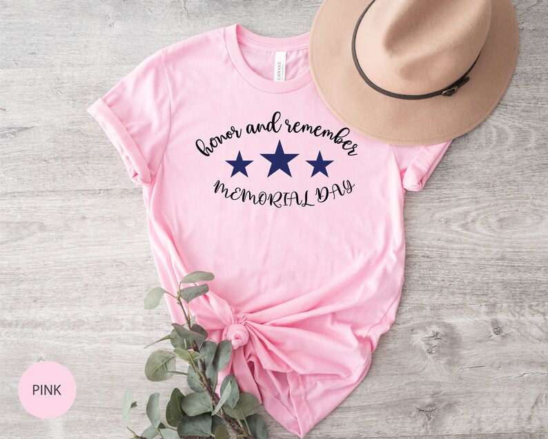 以下が含まれることがあります： 「honor and remember MEMORIAL DAY」という文字と3つの青い星がプリントされたピンクのTシャツです。