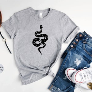 May include: A heather grey t-shirt with a black and white graphic of a snake with stars and moons on its body. The shirt is laid flat on a white surface with a pair of jeans and white sneakers.