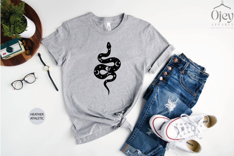 May include: A heather grey t-shirt with a black and white graphic of a snake with stars and moons on its body. The shirt is laid flat on a white surface with a pair of jeans and white sneakers.