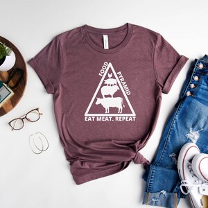 May include: A maroon t-shirt with a white graphic of a food pyramid featuring a cow, pig, and chicken. The text on the shirt reads "Food Pyramid Eat Meat. Repeat."
