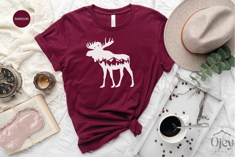 May include: Maroon t-shirt featuring a white moose silhouette with a mountain range design inside. The shirt is displayed on a white surface with a hat, coffee, and other accessories. The shirt is from Ojev Apparel.