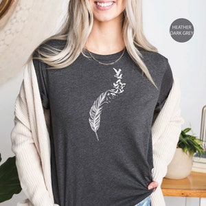 Op de afbeelding: Een donkergrijs heather t-shirt met een witte afbeelding van een veer met vogels die er vanaf vliegen. Het t-shirt wordt gedragen door een persoon met lang blond haar. De tekst "Ojev Apparel" en "Soft Quality Clothing" is zichtbaar in de rechterbovenhoek van de afbeelding. De tekst "Heather Dark Grey" is zichtbaar in de rechterbenedenhoek van de afbeelding.