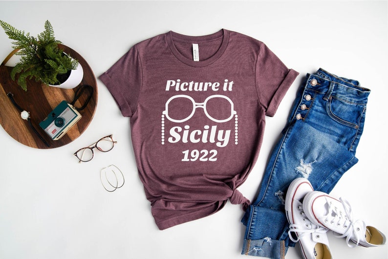 Pu&ograve; includere: Una maglietta color marsala con testo bianco che recita "Picture it Sicily 1922" e una grafica di un paio di occhiali da sole con una collana di perle.