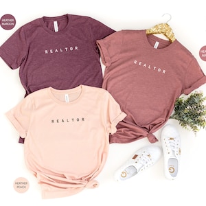 Op de afbeelding: Drie T-shirts met het woord "REALTOR" erop gedrukt. De shirts zijn in de kleuren heather maroon, heather mauve en heather peach.