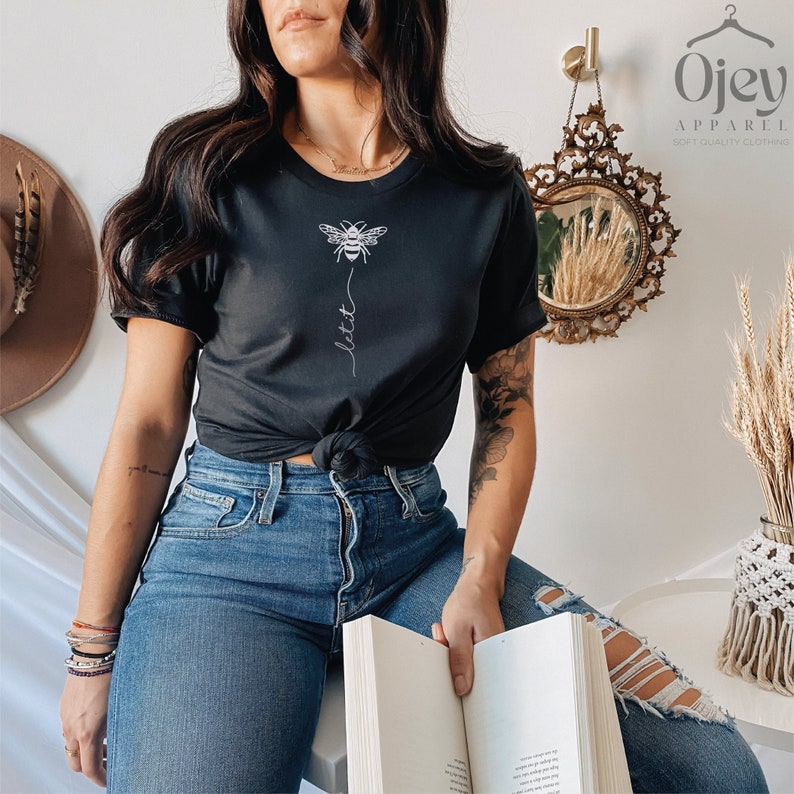 May include: A black t-shirt with a white bee graphic and the word "lett" written vertically below it. The shirt is being worn by a person with a tattoo on their arm.