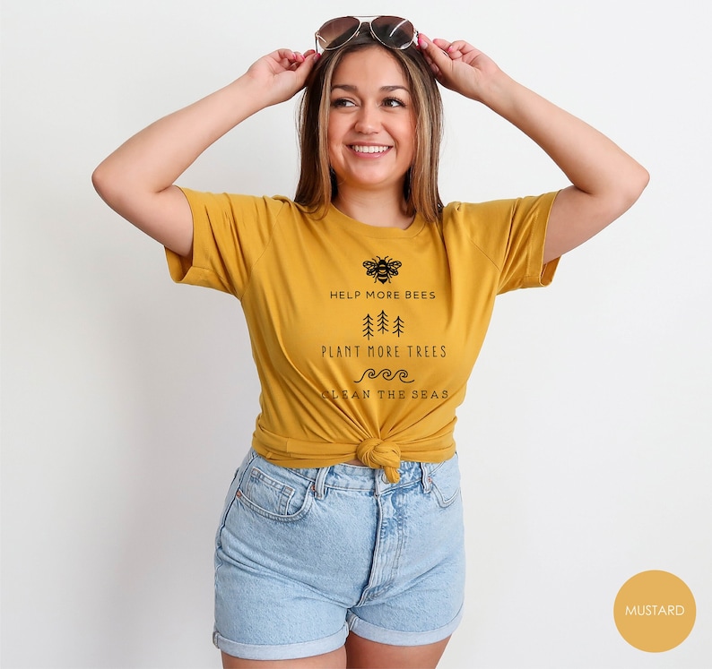K&ouml;nnte beinhalten: Ein senfgelbes T-Shirt mit einer schwarzen Grafik, auf der "Help More Bees, Plant More Trees, Clean the Seas" steht.