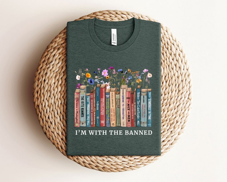 Puede incluir: Una camiseta verde oscuro con un gr&aacute;fico de una estanter&iacute;a llena de libros con t&iacute;tulos como "The Kite Runner", "To Kill a Mockingbird", "The Giver" y "A Wrinkle in Time". El gr&aacute;fico tambi&eacute;n incluye flores y el texto "I'm with the Banned".
