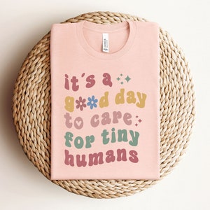 May include: Pink t-shirt with the text "it's a good day to care for tiny humans" in colorful, retro-style lettering.