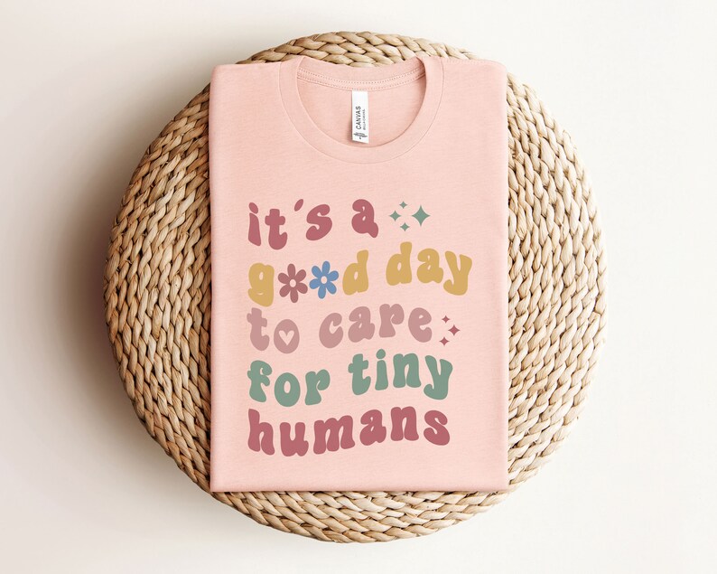 May include: Pink t-shirt with the text "it's a good day to care for tiny humans" in colorful, retro-style lettering.