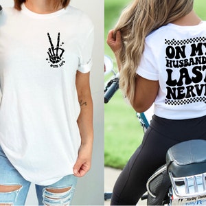 May include: Two white t-shirts with black text and graphics. The first shirt has a skeleton hand making a peace sign with the text "Wife Life". The second shirt has the text "On My Husband's Last Nerve" with a checkered pattern.