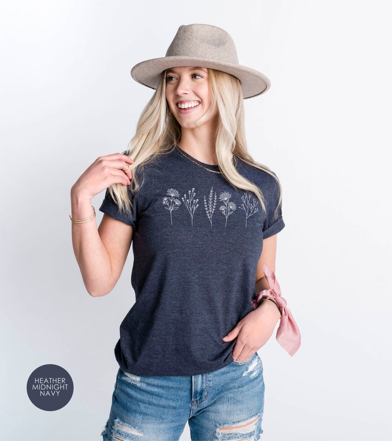 May include: A heather midnight navy t-shirt with a white floral design. The shirt is being worn by a person with blonde hair and a grey hat.