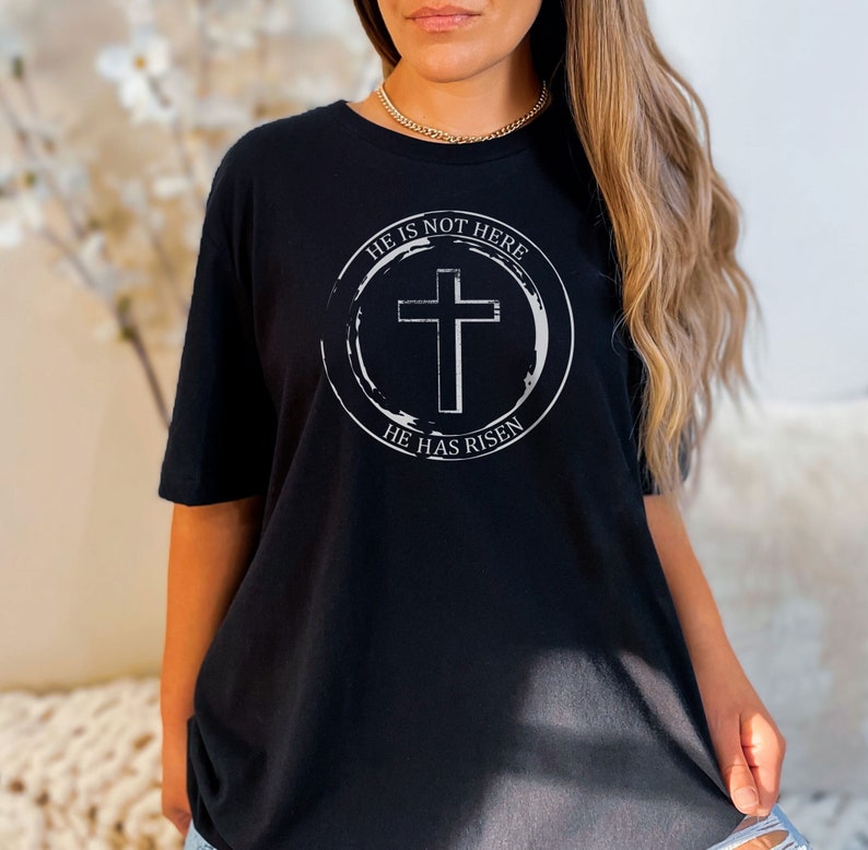 K&ouml;nnte beinhalten: Schwarzes T-Shirt mit einem wei&szlig;en Kreis-Motiv, das ein Kreuz und den Text "Er ist nicht hier, Er ist auferstanden" zeigt.