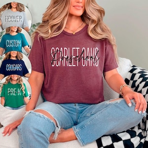 以下が含まれることがあります： 「SCARLET OAKS Preschool」の白文字がプリントされたマルーン色のTシャツ。画像には、「HOOVER」、「CUSTOM」、「COUGARS」、「PRE-K」の文字が入った他の4枚のTシャツも写っています。人物は青いジーンズを着用しています。