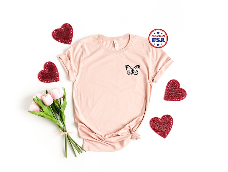 May include: A light pink t-shirt with a black butterfly graphic on the left chest. The shirt is surrounded by red hearts and a bouquet of pink tulips. The shirt has the text "Made in USA" on a blue and white circle.