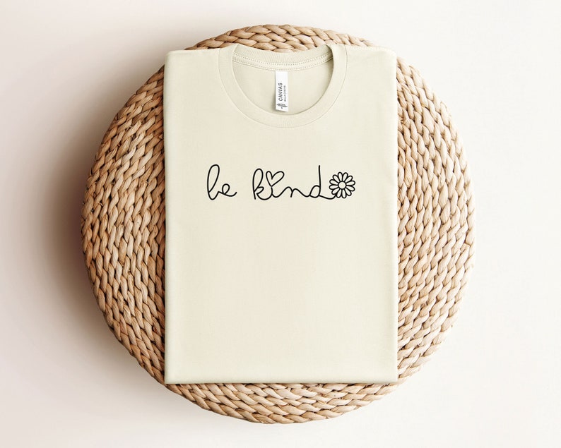 Puede incluir: Una camiseta beige con el texto "be kind" en letra negra con un peque&ntilde;o coraz&oacute;n y un gr&aacute;fico de margarita.