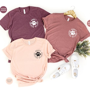 May include: Three t-shirts with the text "Adopt-Foster-Rescue" and a paw print in a circle. The shirts are in heather maroon, heather peach, and heather mauve.