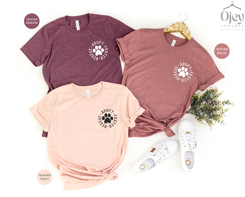 May include: Three t-shirts with the text "Adopt-Foster-Rescue" and a paw print in a circle. The shirts are in heather maroon, heather peach, and heather mauve.