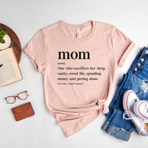 Może przedstawiać: R&oacute;żowy T-shirt z napisem "mom" zdefiniowanym jako "Osoba, kt&oacute;ra poświęca sw&oacute;j sen, zdrowie psychiczne, życie towarzyskie, pieniądze i kt&oacute;ra sika sama (patrz także 'superwoman')"