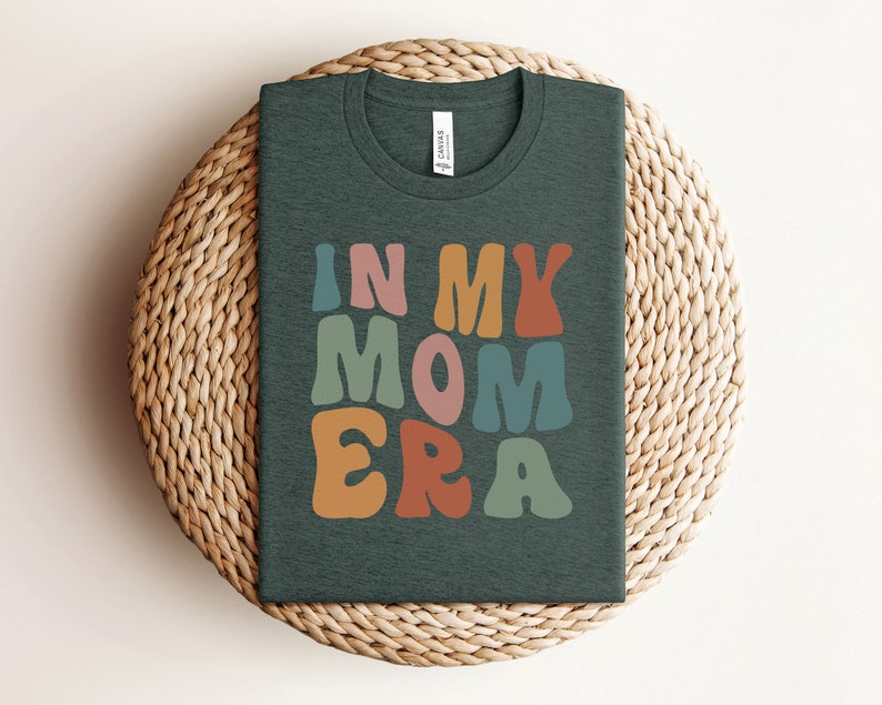May include: A dark green t-shirt with the text "In My Mom Era" in colorful, retro-style lettering.