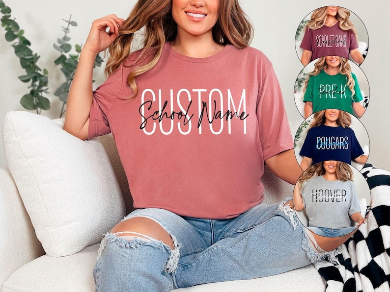 以下が含まれることがあります： 白と黒の筆記体で「CUSTOM School Name」と書かれたダスティローズのTシャツ。画像には「Scarlet Oaks」、「Pre-K」、「Cougars」、「Hoover」と書かれたTシャツも表示されています。Tシャツはカジュアルで、柔らかい素材でできているようです。