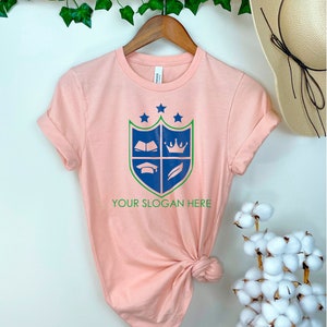 K&ouml;nnte beinhalten: Ein pfirsichfarbenes T-Shirt mit einem blauen und gr&uuml;nen Wappen, das ein Buch, eine Krone, eine Graduation Cap und eine Feder zeigt. Der Text "YOUR SLOGAN HERE" ist unter dem Wappen gedruckt.