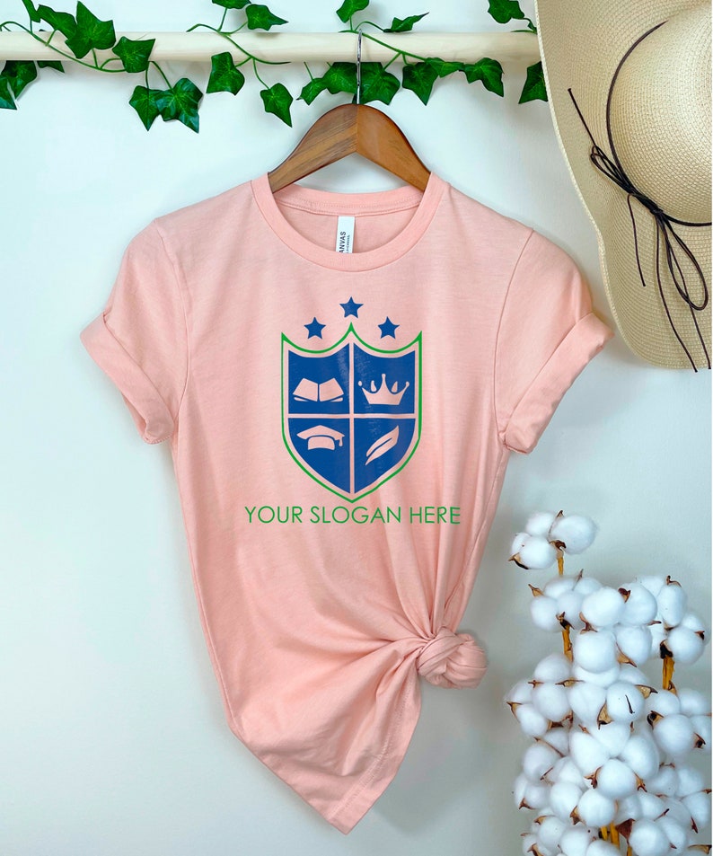 K&ouml;nnte beinhalten: Ein pfirsichfarbenes T-Shirt mit einem blauen und gr&uuml;nen Wappen, das ein Buch, eine Krone, eine Graduation Cap und eine Feder zeigt. Der Text "YOUR SLOGAN HERE" ist unter dem Wappen gedruckt.