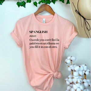 Puede incluir: Una camiseta rosa con la palabra "Spanglish" definida como "Cuando you can't find la palabra en un idioma so you fill it in con el otro."