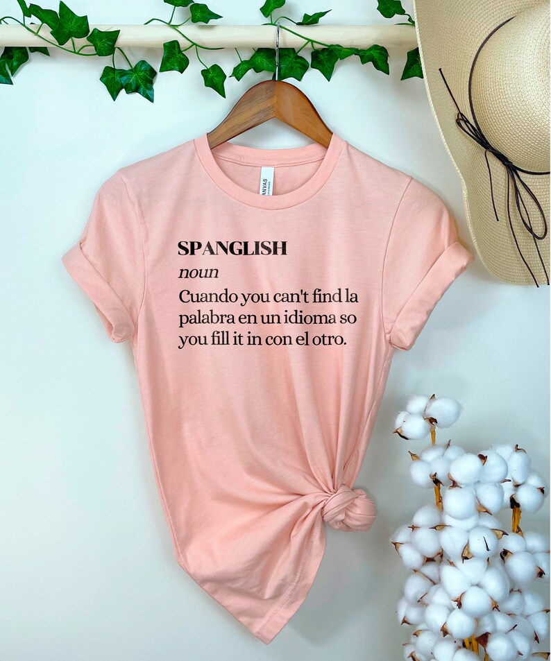 Puede incluir: Una camiseta rosa con la palabra "Spanglish" definida como "Cuando you can't find la palabra en un idioma so you fill it in con el otro."