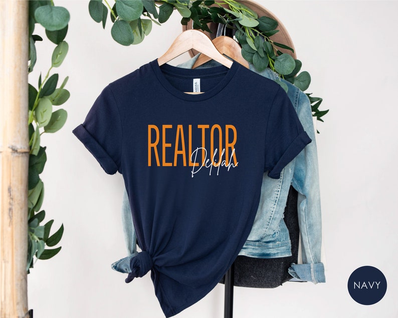 Peut inclure: Un t-shirt bleu marine avec le texte "REALTOR" en orange et "Dejah" en blanc.