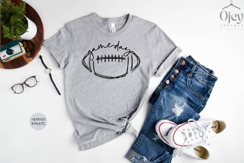 Peut inclure: Un t-shirt gris chin&eacute; avec un graphique noir d'un ballon de football et le texte "game day".
