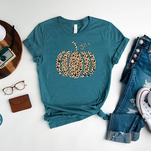 May include: A teal blue t-shirt with a leopard print pumpkin graphic. The shirt is made of a soft, comfortable material and is perfect for fall.