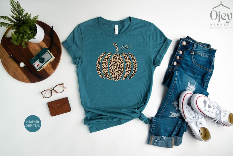 May include: A teal blue t-shirt with a leopard print pumpkin graphic. The shirt is made of a soft, comfortable material and is perfect for fall.