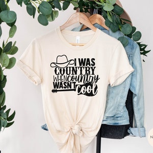 May include: A beige t-shirt with a black graphic that reads "I was country when country wasn't cool". The graphic includes a cowboy hat and a guitar.