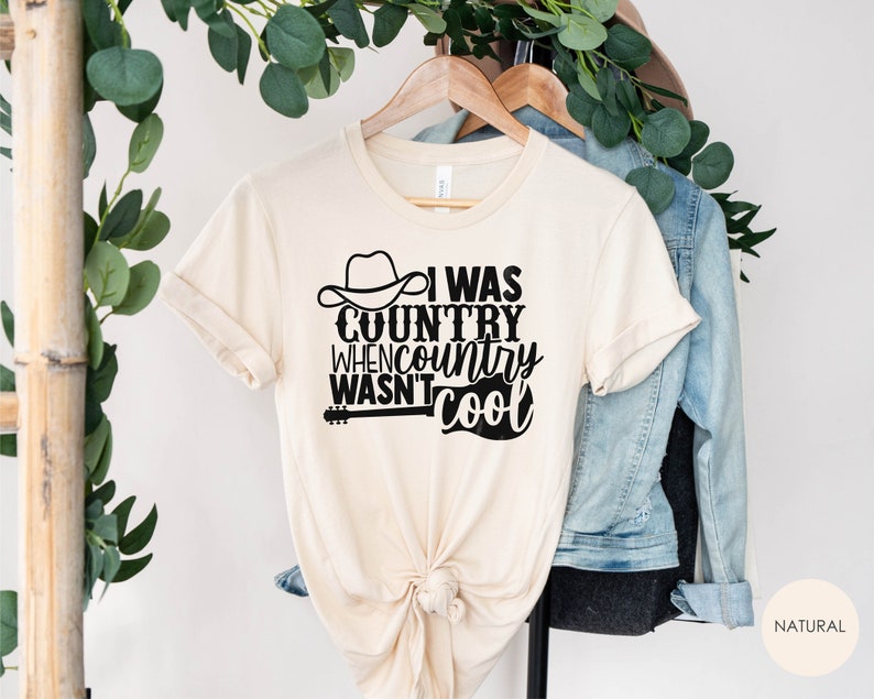 May include: A beige t-shirt with a black graphic that reads "I was country when country wasn't cool". The graphic includes a cowboy hat and a guitar.