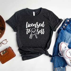 Pode incluir: Camiseta cinza escuro heather com o texto "Licensed to Sell" e um gr&aacute;fico de duas chaves com a hashtag "#realtor" em uma das chaves.