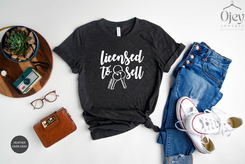 Pode incluir: Camiseta cinza escuro heather com o texto "Licensed to Sell" e um gr&aacute;fico de duas chaves com a hashtag "#realtor" em uma das chaves.