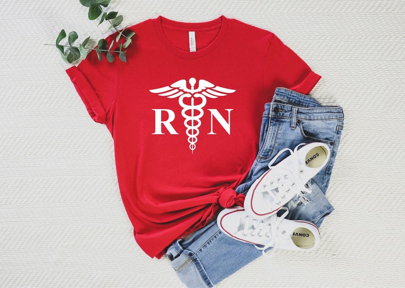 May include: Red t-shirt with a white medical symbol and the letters "RN" printed on the front. The shirt is folded and laid on a white surface with a pair of blue jeans and white sneakers.
