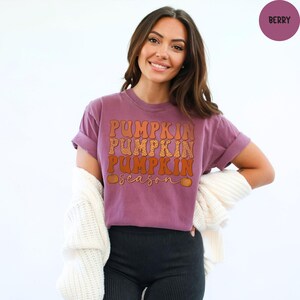 May include: A berry-colored t-shirt with the words "Pumpkin Season" in orange and brown lettering. The shirt has rolled-up sleeves and is being worn with black pants and a white cardigan.