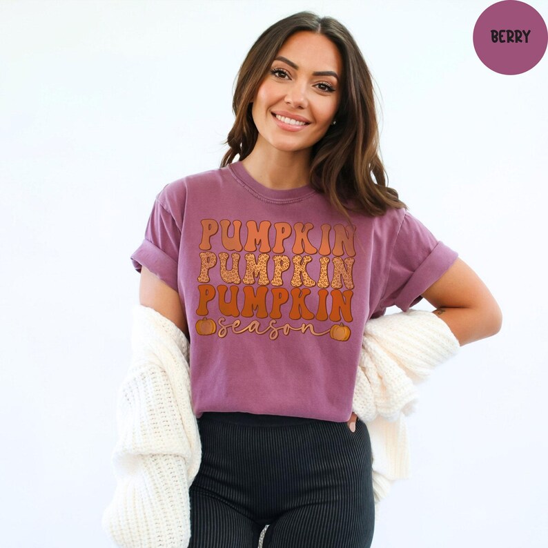 May include: A berry-colored t-shirt with the words "Pumpkin Season" in orange and brown lettering. The shirt has rolled-up sleeves and is being worn with black pants and a white cardigan.