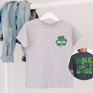K&ouml;nnte beinhalten: Graues T-Shirt mit einem gr&uuml;nen Kleeblatt und dem Namen "MILLER" auf der Vorderseite. Ein dunkelgraues T-Shirt mit einem gr&uuml;nen Kleeblatt und dem Text "ONE LUCKY DUDE" auf der R&uuml;ckseite.