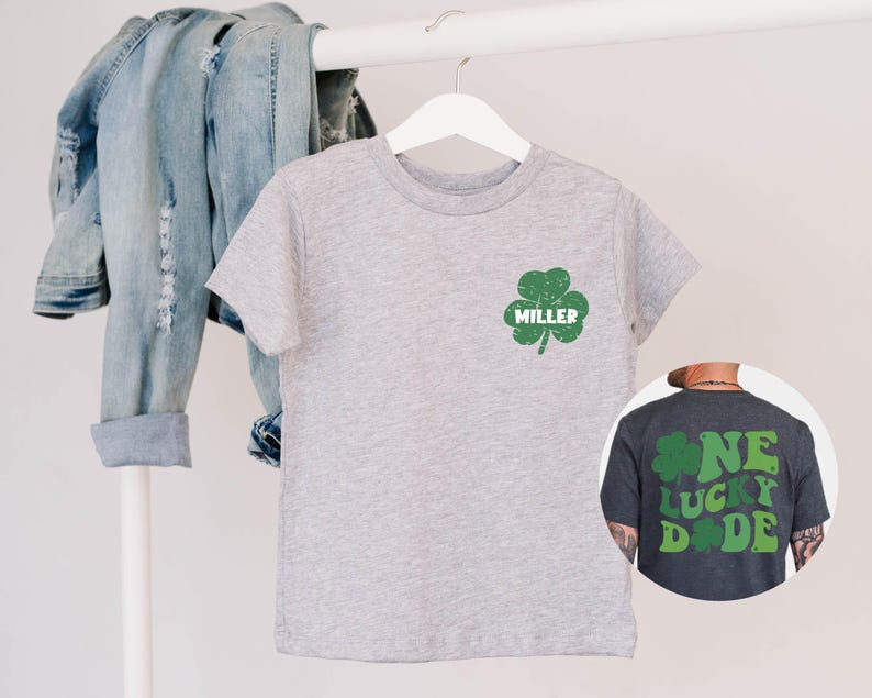 K&ouml;nnte beinhalten: Graues T-Shirt mit einem gr&uuml;nen Kleeblatt und dem Namen "MILLER" auf der Vorderseite. Ein dunkelgraues T-Shirt mit einem gr&uuml;nen Kleeblatt und dem Text "ONE LUCKY DUDE" auf der R&uuml;ckseite.