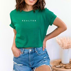 Op de afbeelding: Een vrouw draagt een groen T-shirt met het woord "REALTOR" erop gedrukt. Ze draagt een blauwe denim short en heeft haar handen in haar zakken.