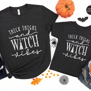 May include: Two heather dark grey t-shirts with the text "Thick Thighs and Witch Vibes" printed in white. The shirts are shown on a white background with fall-themed decorations.