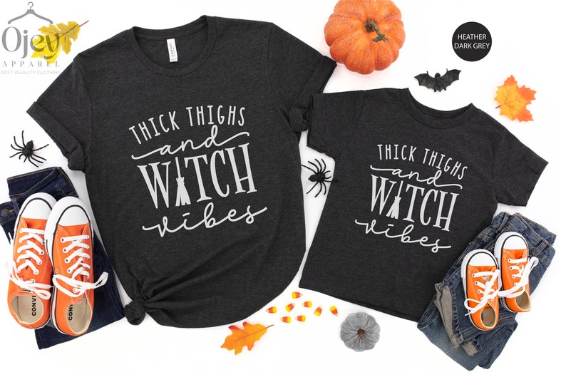 May include: Two heather dark grey t-shirts with the text "Thick Thighs and Witch Vibes" printed in white. The shirts are shown on a white background with fall-themed decorations.