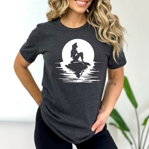 K&ouml;nnte beinhalten: Ein dunkelgraues T-Shirt mit einer wei&szlig;en Silhouette einer Meerjungfrau, die auf einem Felsen vor einem Vollmond sitzt. Der Mond spiegelt sich im Wasser darunter.