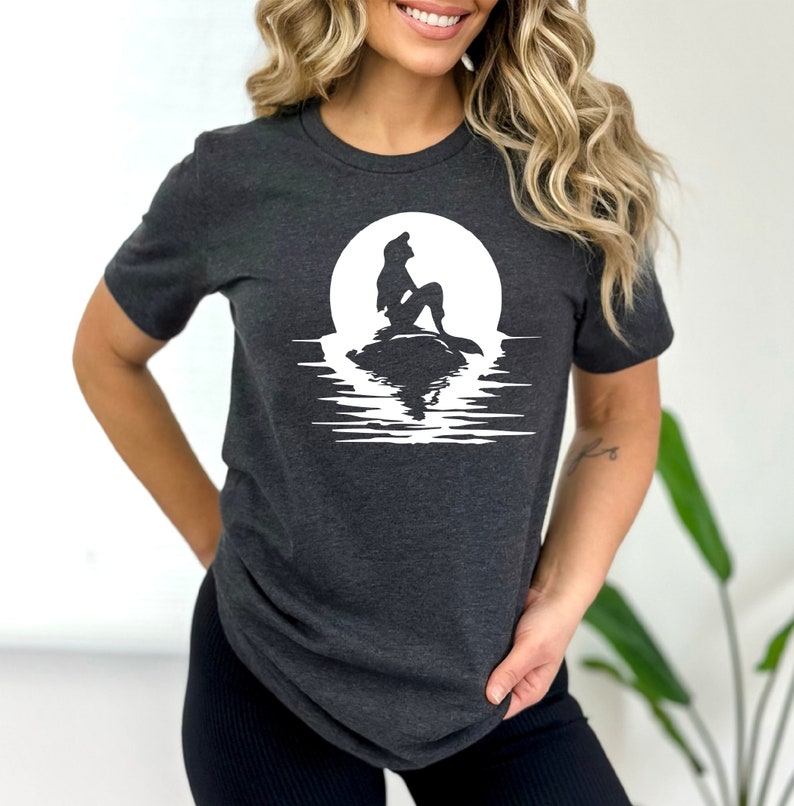 K&ouml;nnte beinhalten: Ein dunkelgraues T-Shirt mit einer wei&szlig;en Silhouette einer Meerjungfrau, die auf einem Felsen vor einem Vollmond sitzt. Der Mond spiegelt sich im Wasser darunter.