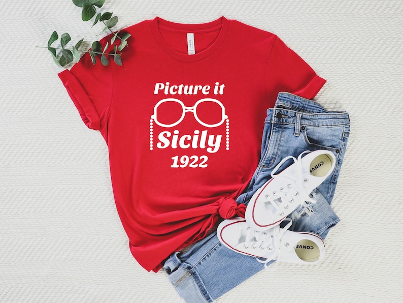 Pu&ograve; includere: Maglietta rossa con testo bianco che recita "Picture it Sicily 1922" e una grafica di occhiali da sole con una catena.