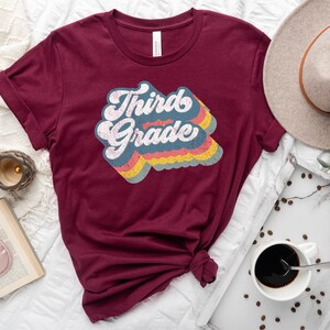 May include: A maroon t-shirt with a retro-style graphic that says "Third Grade" in rainbow colours.