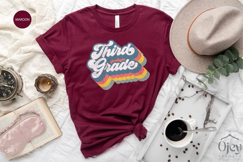 May include: A maroon t-shirt with a retro-style graphic that says "Third Grade" in rainbow colours.